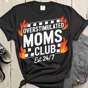 May include: Black t-shirt with the text "OVERSTIMULATED MOMS CLUB Est. 24/7" in white, with flames on either side. The shirt is knotted at the bottom. Black and white sneakers are visible on the left.