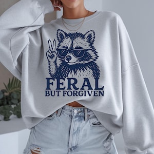 May include: A light gray sweatshirt featuring a navy blue raccoon wearing sunglasses and giving a peace sign. The text "FERAL BUT FORGIVEN" is printed below the raccoon in navy blue.