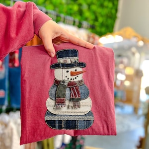 May include: A red t-shirt with a snowman applique. The snowman has a denim hat and body, a carrot nose, and a patterned scarf. The shirt is being held up by a person.