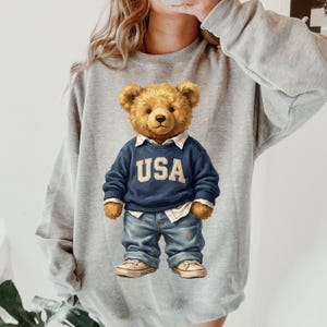 May include: A gray sweatshirt featuring a teddy bear wearing a navy blue sweater with "USA" in white letters, blue jeans, and sneakers. The bear has a light brown fur and is wearing a white collared shirt underneath the sweater.