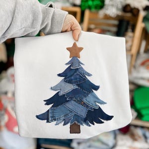Peut inclure: Sweat-shirt blanc orné d'un motif de sapin de Noël. L'arbre est composé de différentes nuances de denim bleu, avec une étoile et un tronc marron. Le sweat-shirt est tenu par une main.