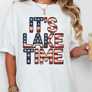 以下が含まれることがあります： アメリカの旗のデザインで、赤、白、青の「IT'S LAKE TIME」の文字が書かれた白いTシャツ。