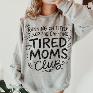 May include: A light gray sweatshirt with the text "RUNNING ON LITTLE SLEEP AND CAFFEINE TIRED MOMS CLUB" in black. The design includes a coffee cup, lightning bolts, and decorative swirls. The sweatshirt is a casual style.