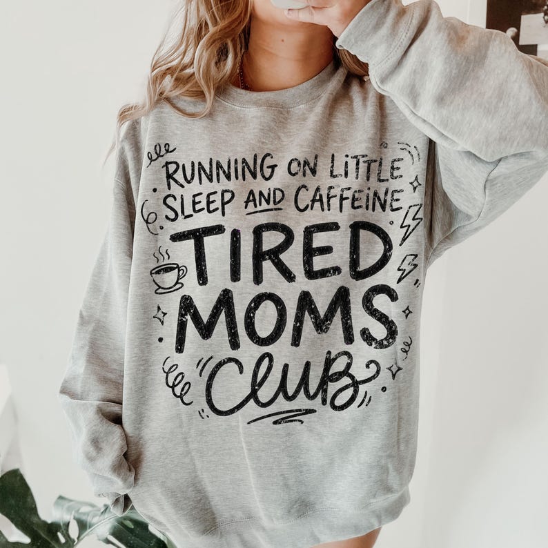 May include: A light gray sweatshirt with the text "RUNNING ON LITTLE SLEEP AND CAFFEINE TIRED MOMS CLUB" in black. The design includes a coffee cup, lightning bolts, and decorative swirls. The sweatshirt is a casual style.