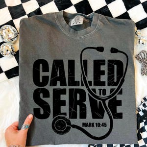 Puede incluir: Camiseta gris con las palabras "CALLED TO SERVE" en letras negras grandes, con un gráfico de estetoscopio. El texto "MARK 10:45" está debajo del estetoscopio. La camiseta se muestra sobre una superficie blanca con tela a cuadros en blanco y negro.
