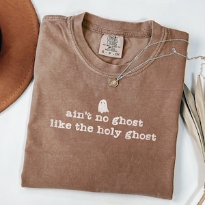 May include: A brown Comfort Colors t-shirt with the phrase "ain't no ghost like the holy ghost" and a ghost graphic. A silver necklace with a pendant is draped across the shirt. A brown hat is visible in the upper left corner.