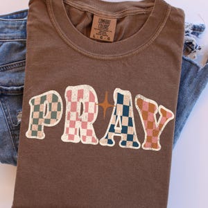 May include: A brown t-shirt with the word "PRAY" in a retro font. The letters are filled with checkered patterns in shades of pink, blue, and brown. A star graphic is between the "R" and "A". The shirt is paired with blue jeans.