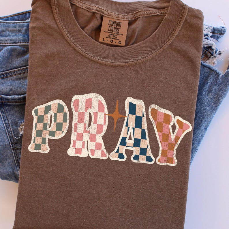 May include: A brown t-shirt with the word "PRAY" in a retro font. The letters are filled with checkered patterns in shades of pink, blue, and brown. A star graphic is between the "R" and "A". The shirt is paired with blue jeans.