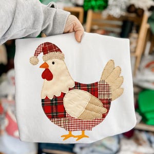 May include: White sweatshirt featuring an appliqued chicken wearing a Santa hat. The chicken has a red wattle and beak, with a plaid pattern on its body and hat. The design is stitched onto the sweatshirt.