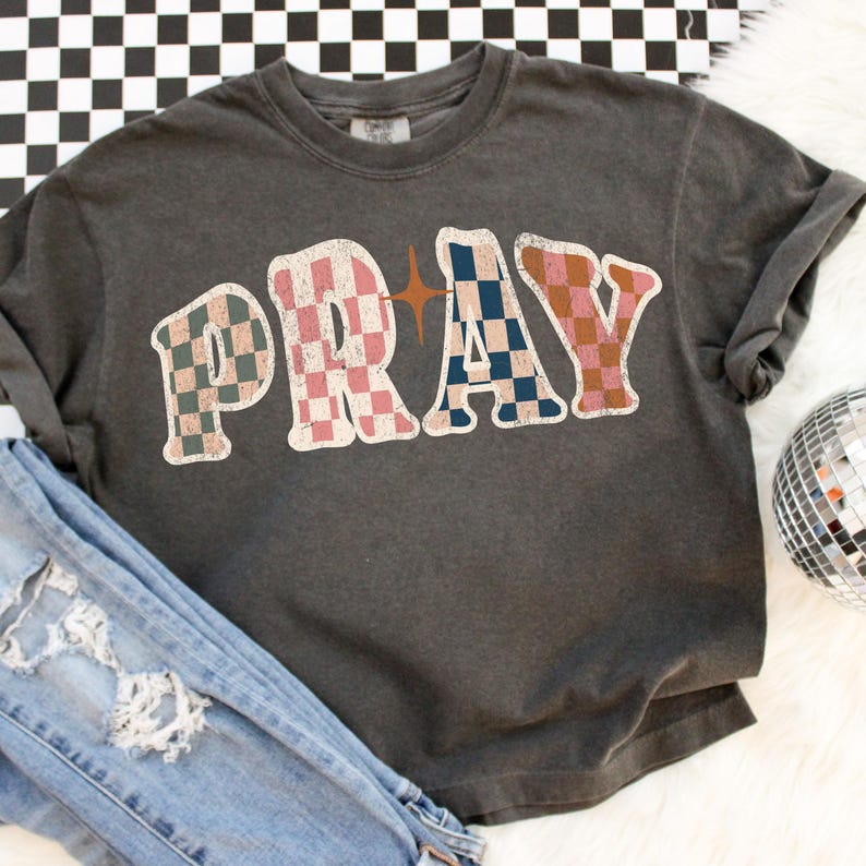 May include: A dark gray t-shirt with the word "PRAY" in a retro, checkered design. The letters are in pink, blue, and brown, with a star graphic. The shirt is paired with distressed denim jeans and a disco ball.