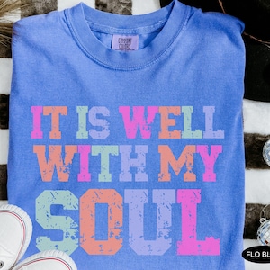 May include: A blue Comfort Colors t-shirt with the phrase "IT IS WELL WITH MY SOUL" printed in colorful, distressed lettering. The shirt is a solid blue color and has a crew neck. The shirt is paired with white sneakers.