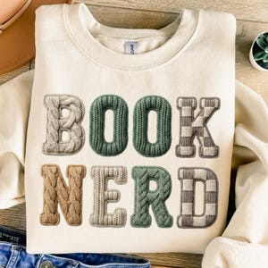 May include: A cream-coloured sweatshirt featuring the text "BOOK NERD" in a textured, knitted design. The letters are in beige, green, and brown, with the "K" and "D" in a checkered pattern. A comfortable, casual top for book enthusiasts.