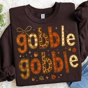May include: A dark brown sweatshirt with the words "Gobble Gobble" in a fall-themed design. The letters feature plaid, leopard print, and dotted patterns, with decorative acorns and leaves. A perfect Thanksgiving or autumn-themed apparel.
