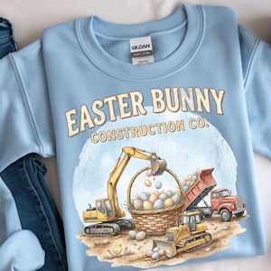 Può includere: Felpa azzurra con la scritta "EASTER BUNNY CONSTRUCTION CO." e una grafica di veicoli da cantiere, un cesto di uova di Pasqua e pulcini. Realizzata in cotone pesante.