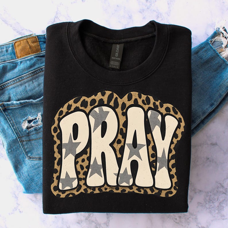 May include: Black crew neck sweatshirt featuring the word "PRAY" in a retro font, filled with grey stars. The word is outlined with a leopard print design. The sweatshirt is displayed with a pair of blue jeans.