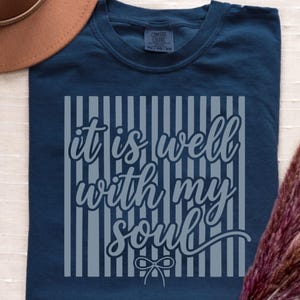 May include: A navy blue t-shirt with the phrase "it is well with my soul" in a light gray font. The text is centered within vertical gray stripes. The shirt is made of a soft material.