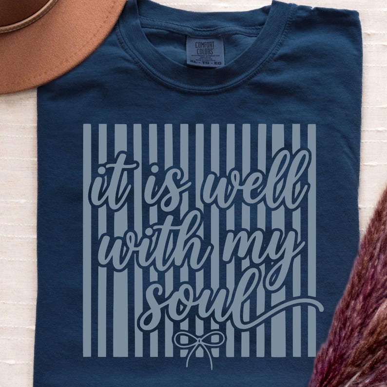 May include: A navy blue t-shirt with the phrase "it is well with my soul" in a light gray font. The text is centered within vertical gray stripes. The shirt is made of a soft material.