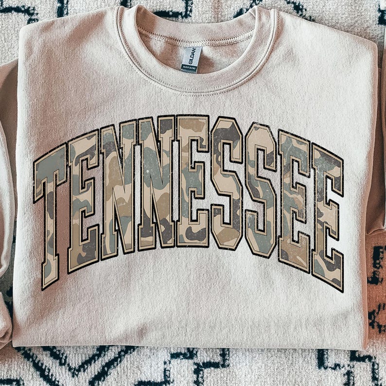 May include: A beige sweatshirt featuring the word "TENNESSEE" in large, block letters. The letters are filled with a camouflage pattern in green, brown, and grey, and are outlined in a darker grey.