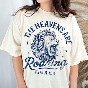 May include: Cream-colored t-shirt with a distressed blue graphic. The graphic features a roaring lion's head, surrounded by the words "THE HEAVENS ARE ROARING" and "PSALM 19:1". The design includes star accents and a vintage aesthetic.
