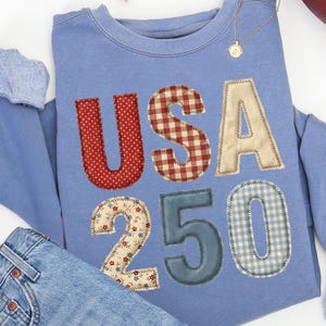 May include: A light blue sweatshirt with the letters "USA" and the numbers "250" appliquéd on the front. The letters and numbers are made of various patterned fabrics, including red polka dots, gingham, and floral prints. A gold necklace is visible.