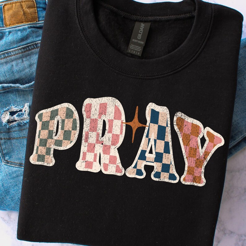 May include: Black sweatshirt with the word "PRAY" in a retro font. The letters are checkered in shades of pink, blue, green, and brown. A gold star graphic is between the "R" and "A". The sweatshirt is folded on top of a pair of blue jeans.