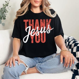 Könnte beinhalten: Schwarzes T-Shirt mit den Worten "THANK YOU Jesus" in roter und weißer Schrift. Das Shirt hat einen Used-Look im Vintage-Stil. Das Model trägt eine blaue Jeans.