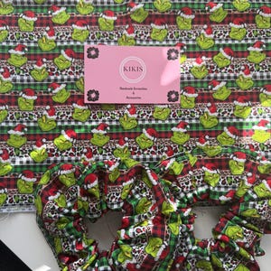 May include: Two fabric scrunchies and a sheet of fabric featuring a repeating pattern of green characters wearing red Santa hats, interspersed with horizontal stripes of black and white buffalo plaid and brown leopard print, with a pink KIKIS card.