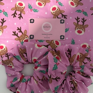 May include: Two pink fabric hair scrunchies featuring a repeating pattern of brown reindeer with red noses and green holly leaves. A rectangular pink business card is centred above the scrunchies with the text KIKIS Handmade Scrunchies & Accessories.