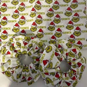 May include: Two fabric hair scrunchies and a matching rectangular fabric piece featuring an all-over print of the Grinch wearing a red Santa hat, interspersed with the word Grinch in green text against a white background.