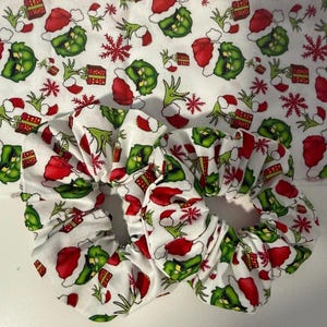 May include: Two fabric hair scrunchies featuring a repeating pattern of green cartoon characters wearing red Santa hats, red gift boxes, and red snowflakes on a white background. The same patterned fabric is visible behind the scrunchies.