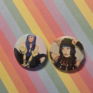 Puede incluir: Dos insignias redondas con imágenes de una persona con cabello azul. Una insignia presenta a una persona con cabello azul y un atuendo amarillo y negro. La otra insignia presenta a una persona con cabello azul y un sombrero negro.