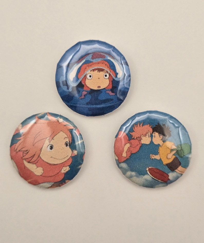 May include: Three round buttons featuring anime characters. One button shows a girl in a red hood in a blue background. Another shows a girl with red hair. The third button shows two characters kissing in a blue sky.