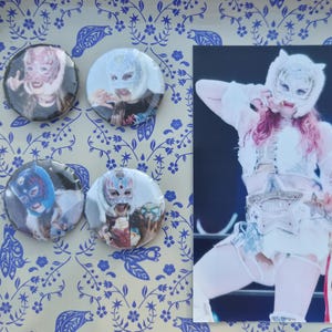 May include: An image featuring four round pinback buttons with luchador mask designs, alongside a photograph of a wrestler in a white and silver costume. The buttons display various colorful mask designs. The wrestler in the photo is wearing a white mask and costume.