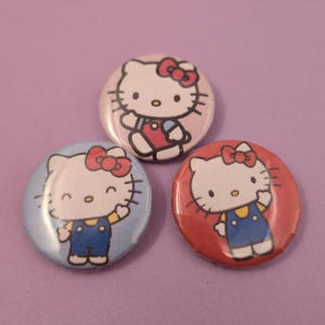 Puede incluir: Tres botones redondos de Hello Kitty. Cada botón presenta un gato blanco con un lazo rojo, con un peto azul. Los botones están sobre un fondo morado, con uno sobre un fondo rosa, uno sobre un fondo azul y otro sobre un fondo rojo.