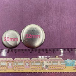 Op de afbeelding: Twee zilverkleurige metalen knopen, &eacute;&eacute;n van 25 mm en de andere van 32 mm in diameter, met roze labels. Onderaan staat een liniaal met een treindesign. De achtergrond is effen paars.