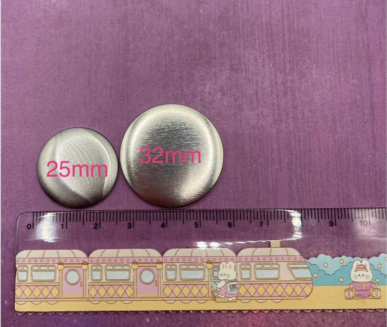 Op de afbeelding: Twee zilverkleurige metalen knopen, &eacute;&eacute;n van 25 mm en de andere van 32 mm in diameter, met roze labels. Onderaan staat een liniaal met een treindesign. De achtergrond is effen paars.