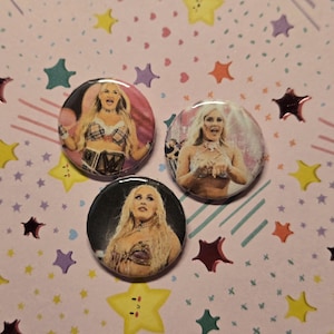 May include: Three round pinback buttons featuring images of a blonde woman in various outfits. The buttons have a glossy finish and are set against a pink background decorated with colorful star and heart shapes.