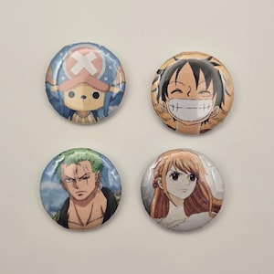 May include: Four round pinback buttons featuring characters from the anime series One Piece. The buttons display images of Tony Tony Chopper, Monkey D. Luffy, Roronoa Zoro, and Nami. Each button has a glossy finish and a pin on the back.