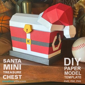 May include: A red and white papercraft Santa Claus treasure chest with a black belt and gold buckle. The chest is open and has a white interior. The chest is sitting on a wooden surface. DIY Paper Model Template SVG, PDF, PNG.