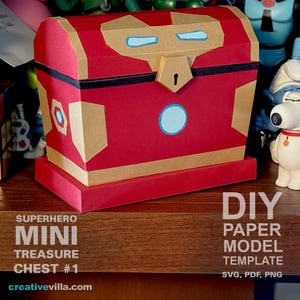 Può includere: Un forziere del tesoro in carta rosso e oro con un design da supereroe. Il forziere ha una fascia nera attorno alla parte superiore e un buco della serratura. Il forziere è appoggiato su una superficie di legno. Modello di carta fai-da-te SVG, PDF, PNG.