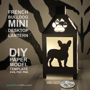 Könnte beinhalten: Eine DIY-Papiermodellvorlage für eine Miniatur-Desktop-Laterne mit einem französischen Bulldoggen. Die Vorlage enthält SVG-, PDF- und PNG-Dateien. Die Laterne ist eine schwarze Silhouette eines französischen Bulldoggen mit einem Pfotenabdruck auf dem Dach. Die Laterne wird von innen beleuchtet und strahlt ein warmes Licht aus.