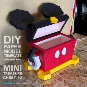 Puede incluir: Un modelo de papel rojo y negro de un cofre del tesoro de Mickey Mouse con una base amarilla y ojos blancos. El cofre está abierto y el interior es rosa. El texto "DIY PAPER MODEL TEMPLATE SVG, PDF, PNG MINI TREASURE CHEST #2 creativevilla.com" está impreso debajo del cofre.