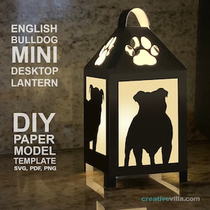 Può includere: Una lanterna di carta nera con una silhouette di Bulldog Inglese su ogni lato. La lanterna ha un manico in cima e ritagli a forma di impronta di zampa. Il testo "ENGLISH BULLDOG MINI DESKTOP LANTERN DIY PAPER MODEL TEMPLATE SVG, PDF, PNG" è stampato sul lato sinistro dell'immagine. La lanterna è illuminata dall'interno, proiettando un bagliore caldo.