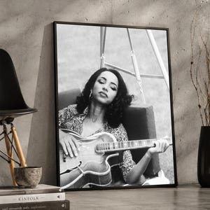 Könnte beinhalten: Ein schwarz-weiß gerahmter Wandkunstdruck, der eine Person mit dunklem Haar zeigt, die auf einer Schaukel sitzt und eine akustische Gitarre spielt. Die Person trägt ein gemustertes Hemd und Creolen, die Augen sind geschlossen.