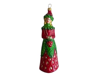 May include: A glass Christmas bauble shaped like a woman wearing a red dress with green leaves and red strawberries. The bauble has a gold loop for hanging.