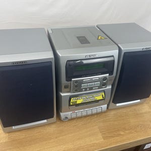 May include: A silver Aiwa stereo system featuring a central unit with a CD player, digital tuner, and cassette deck, flanked by two detachable speakers with dark blue fabric grilles. The central unit displays text regarding power output.