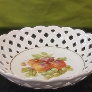 以下が含まれることがあります： 縁に格子模様のある白い磁器製のサービングディッシュ。皿の中央には、赤い果物と黄色の果物、緑の葉とブドウが描かれた静物画が描かれています。金のトリムが縁を強調しています。