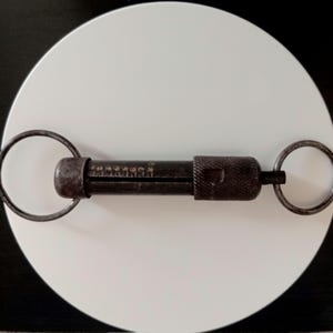 以下が含まれることがあります： 中央のバレルに刻印された数値マーキング、ローレット加工されたグリップセクション、両端に取り付けられた円形の金属リングを備えた、暗い金属製の円筒形ツール。このデバイスは、経年変化が見られる風化した工業的な外観をしています。