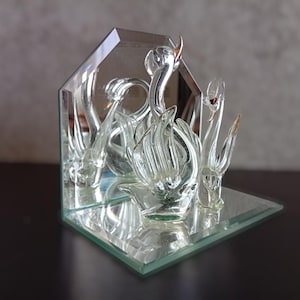 以下が含まれることがあります： 透明なガラスの白鳥の彫刻。3羽の白鳥が鏡面ベースに配置されています。白鳥は繊細なディテールを持ち、鏡面の背景の前に配置され、反射を作り出しています。彫刻は暗い表面に展示されています。