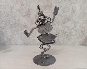 May include: A handcrafted metal sculpture of a dancing figure. The sculpture is made from various metal pieces, including nuts, bolts, and perforated metal. The figure is posed mid-dance, with arms raised and legs bent. The sculpture is mounted on a circular base.